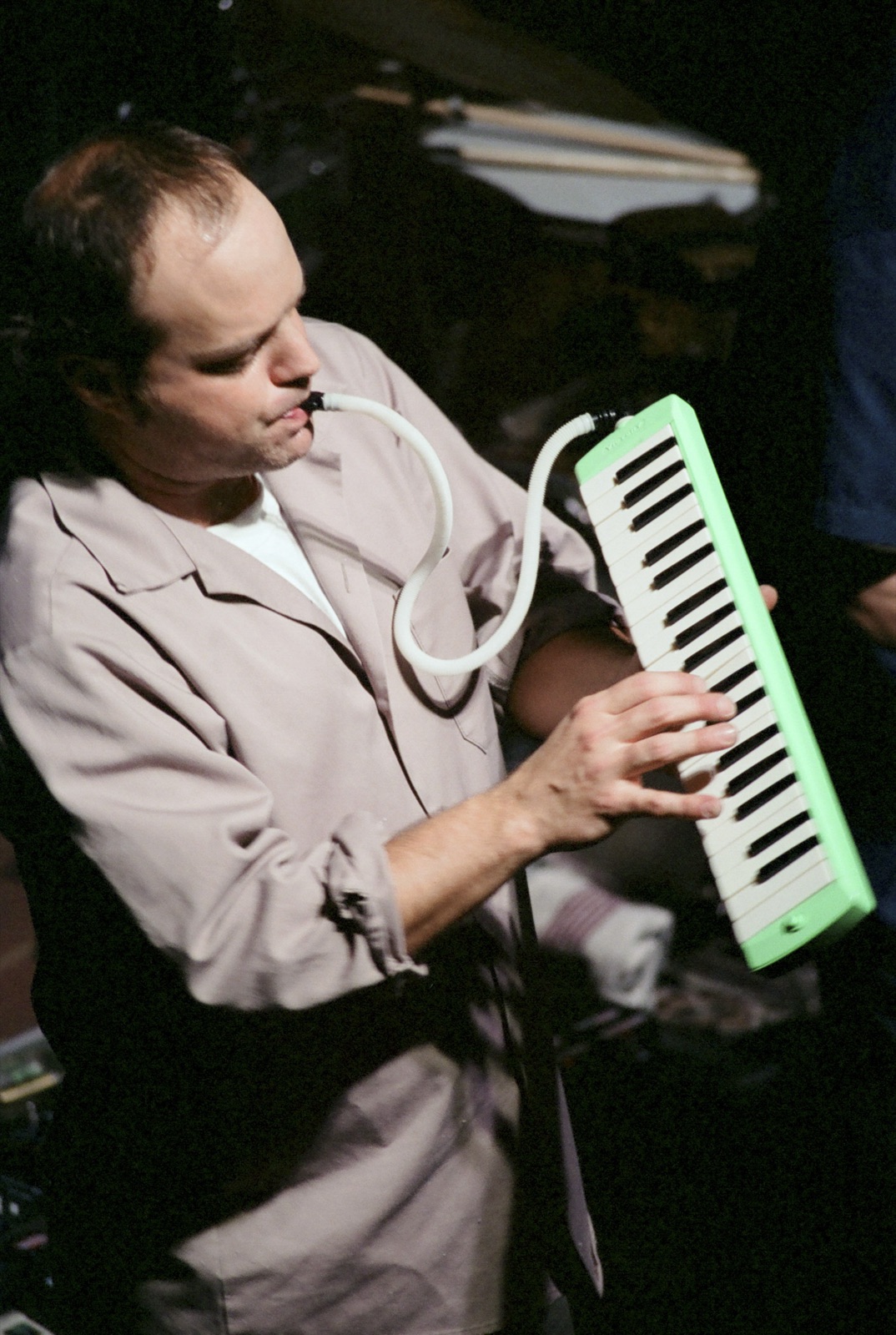 A keyboardist plays a green melodica, its mouthpiece tube looping from his lips to the keys.